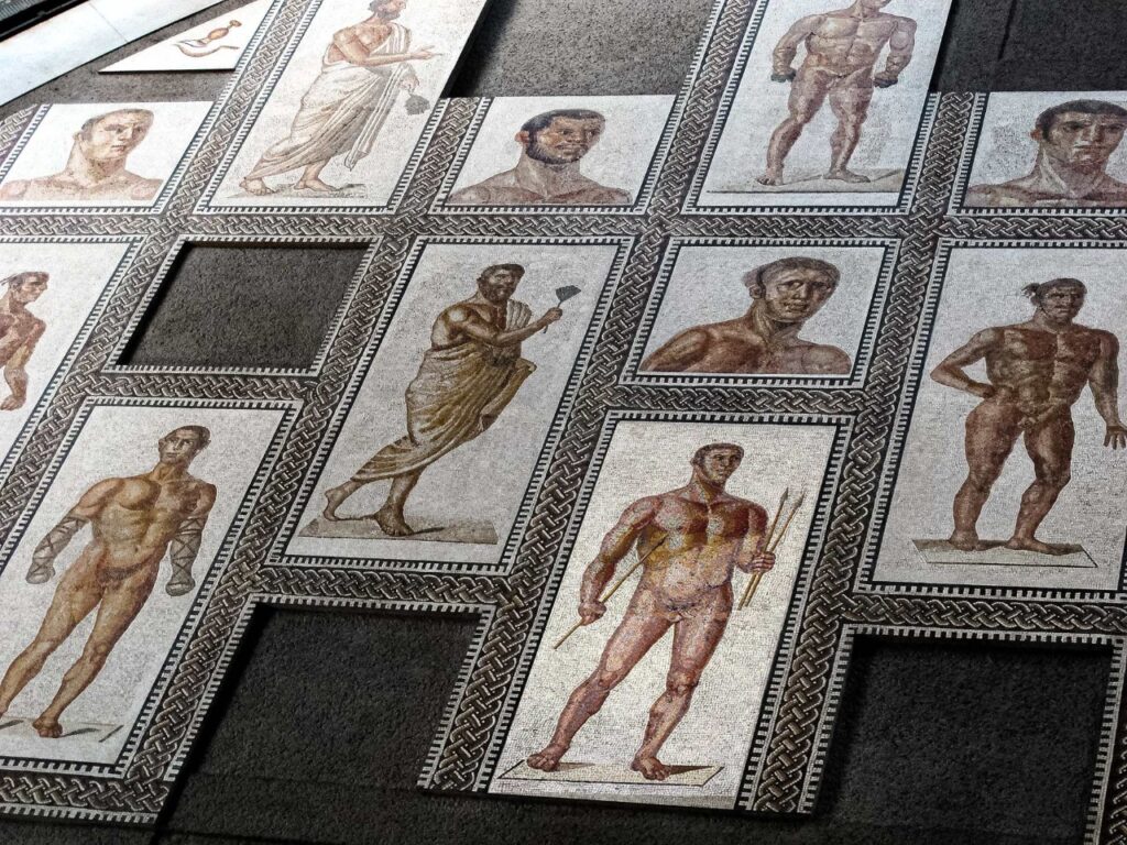 Mosaico dos Banhos de Caracala, representando atletas da Antiguidade em várias poses, com detalhes de armas e posturas esportivas. O estilo clássico é destacado por bordas geométricas e cores vivas. As áreas vazias sugerem restauração ou escavação. Parte importante da arte romana.