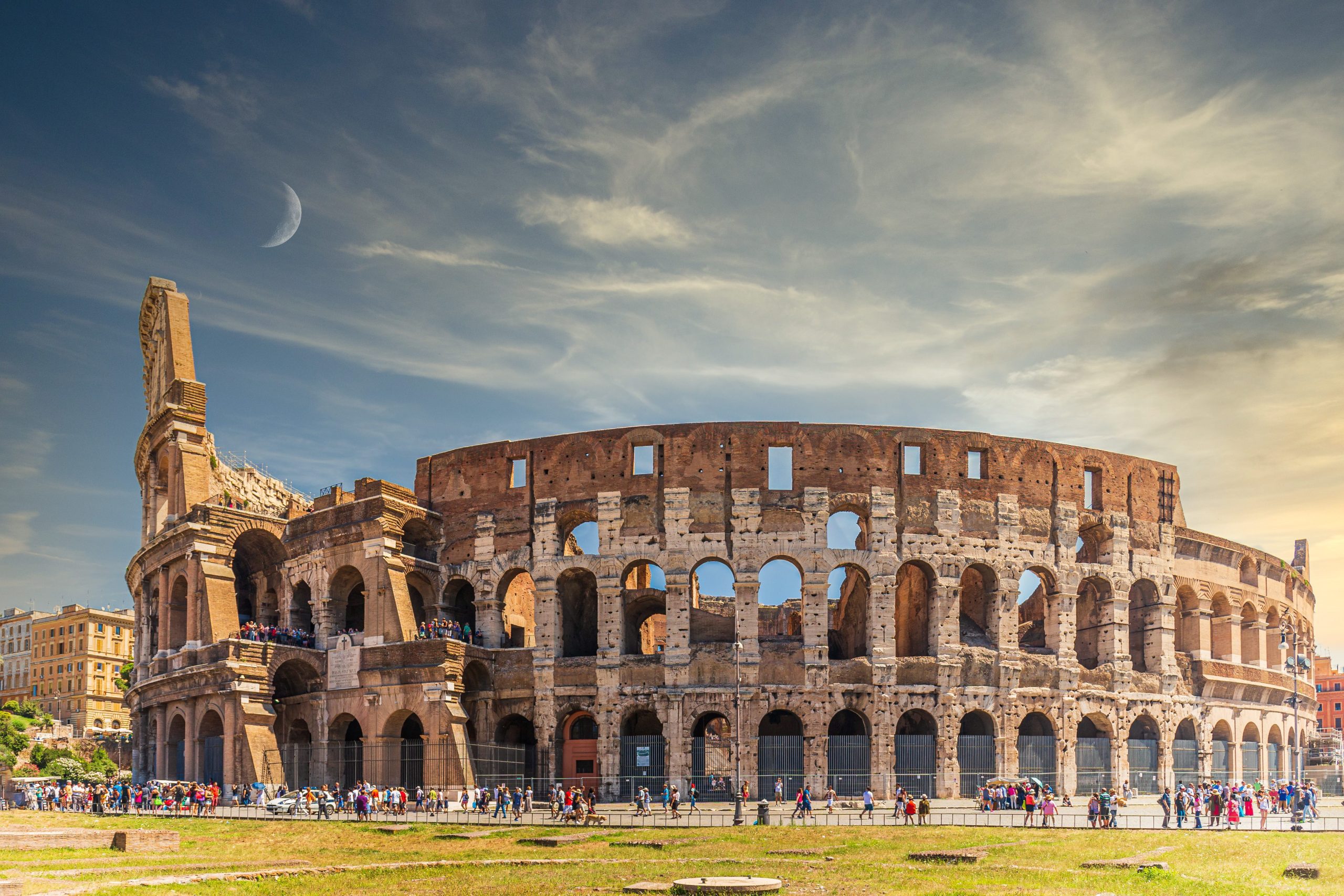 Vista diurna do Coliseu em Roma com turistas ao redor e uma lua crescente visível no céu azul.