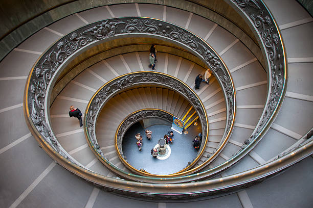 A imagem mostra a famosa Escadaria de Bramante nos Museus Vaticanos. Com seu design em espiral e detalhes ornamentados, a vista de cima destaca suas curvas perfeitas. Pessoas caminham pelas escadas, dando uma sensação de movimento à estrutura elegante. É um ícone da arquitetura renascentista.