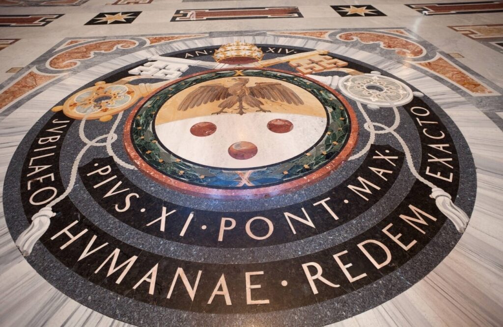 A imagem mostra um detalhe do piso em mármore da Basílica de São Pedro, no Vaticano, com o brasão de Pio XI, incluindo as chaves cruzadas e a tiara papal. O texto em latim celebra o jubileu do Papa Pio XI.