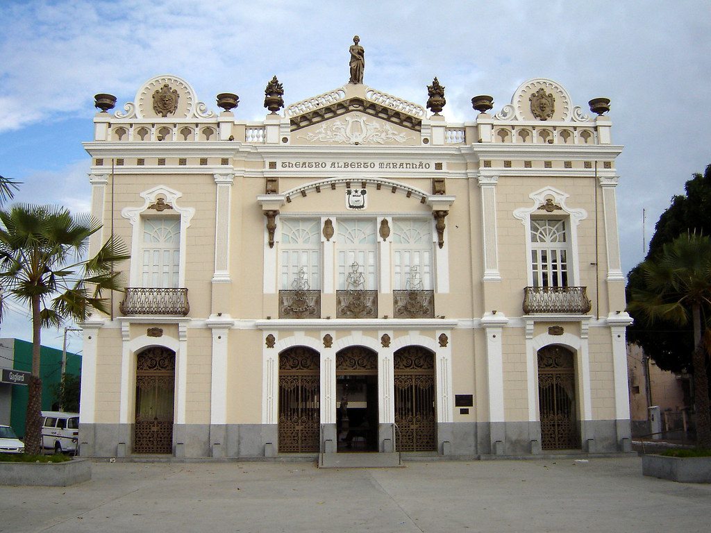 Fachada do Teatro Alberto Maranhão, um edifício histórico com arquitetura eclética, localizado em Natal, Rio Grande do Norte. A construção exibe detalhes ornamentais elaborados e varandas decorativas, com palmeiras ao redor.