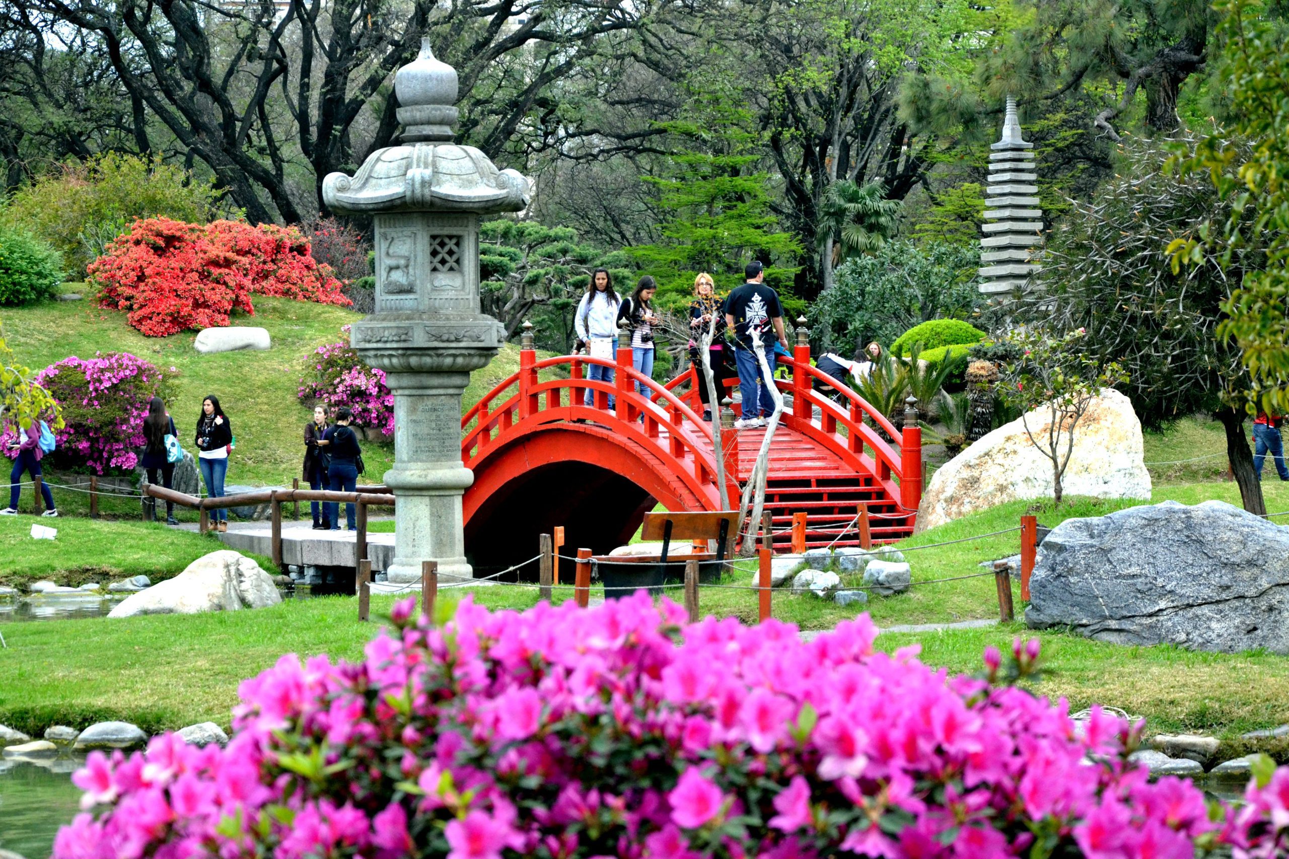 Visitantes atravessam uma ponte vermelha tradicional no Jardim Japonês em Buenos Aires, cercada por flores vibrantes, lanternas de pedra e vegetação exuberante.