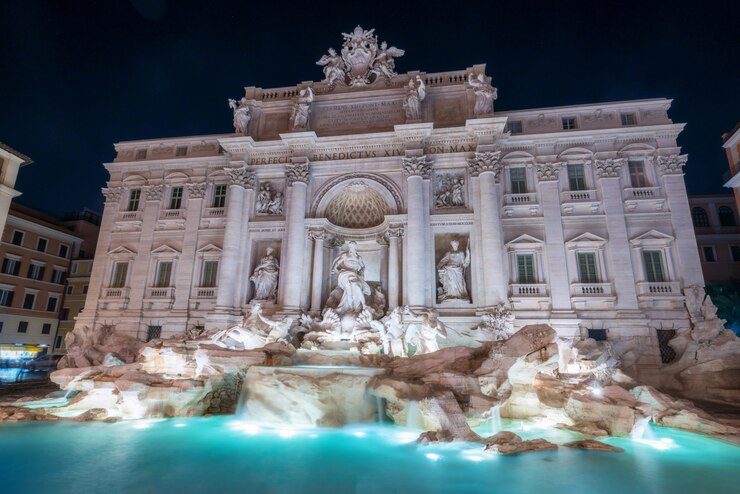 A imagem exibe a majestosa Fontana di Trevi, um dos monumentos mais icônicos de Roma, Itália. À noite, a fonte brilha com iluminação que destaca suas impressionantes esculturas barrocas e a beleza arquitetônica detalhada, criando uma atmosfera mágica e encantadora.