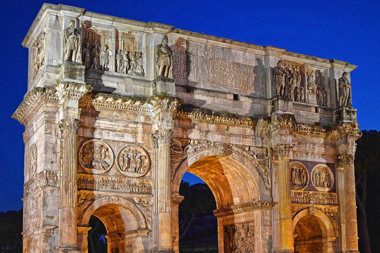 O Arco de Constantino em Roma, iluminado à noite, destaca suas esculturas e relevos detalhados. No topo, uma inscrição central é ladeada por relevos de Marco Aurélio. Abaixo, medalhões da época de Adriano e Constantino e frisos com cenas das vitórias de Constantino.