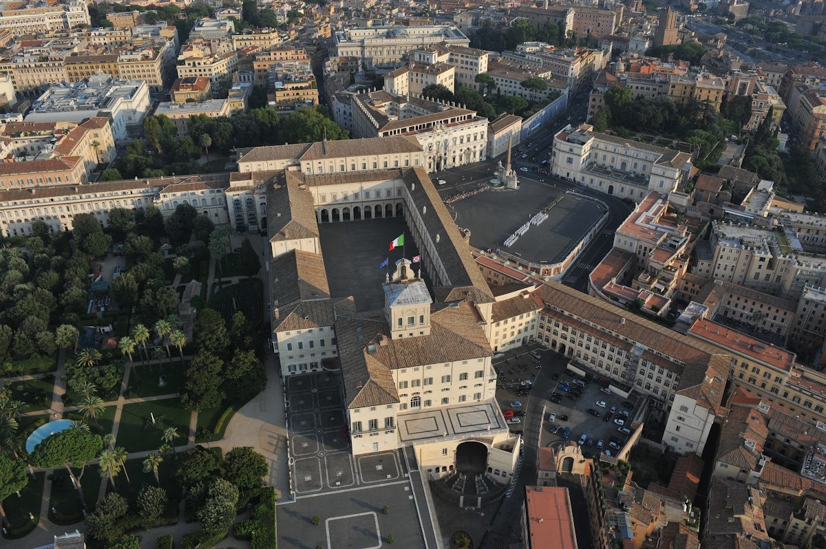 Vista aérea da Colina Quirinal, destacando o imponente Palácio do Quirinal, residência oficial do Presidente da Itália, cercado por jardins luxuriantes e a arquitetura histórica de Roma ao fundo.