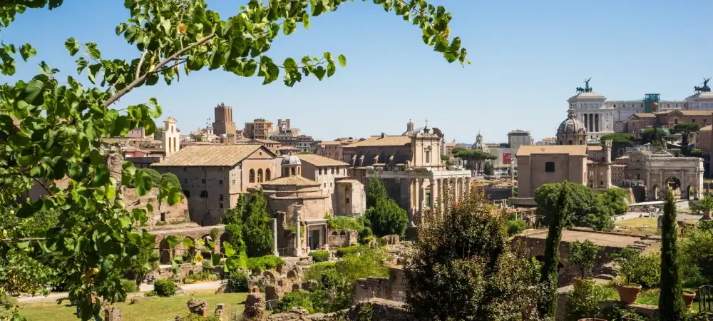 Essa imagem mostra uma vista panorâmica das ruínas antigas de Roma, incluindo diversos templos, arcos e estruturas históricas, cercados por vegetação e com a cidade moderna ao fundo. As ruínas são parte do Fórum Romano, um dos locais mais emblemáticos da história romana.