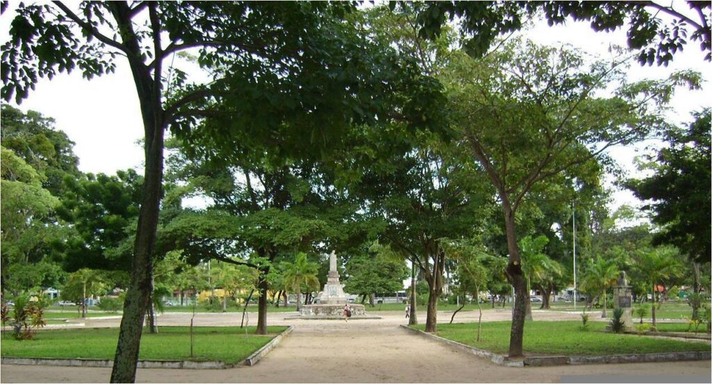 Caminho de terra em uma praça arborizada, ladeado por árvores que formam um túnel natural. Ao fundo, um obelisco é visível, cercado por áreas verdes e outras plantas. O ambiente transmite tranquilidade em meio à vegetação densa.