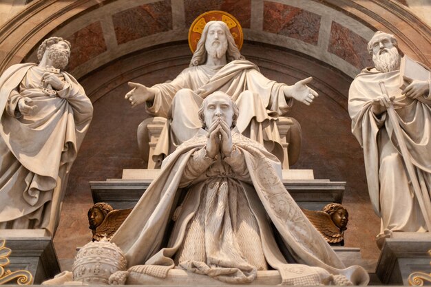 Escultura detalhada dentro da Basílica de São Pedro, no Vaticano, retratando figuras religiosas em oração. No centro, um papa ajoelhado com as mãos unidas em oração, rodeado por estátuas de Cristo e santos em posição de bênção. O cenário é majestoso, com detalhes arquitetônicos e de vestimenta esculpidos em mármore.