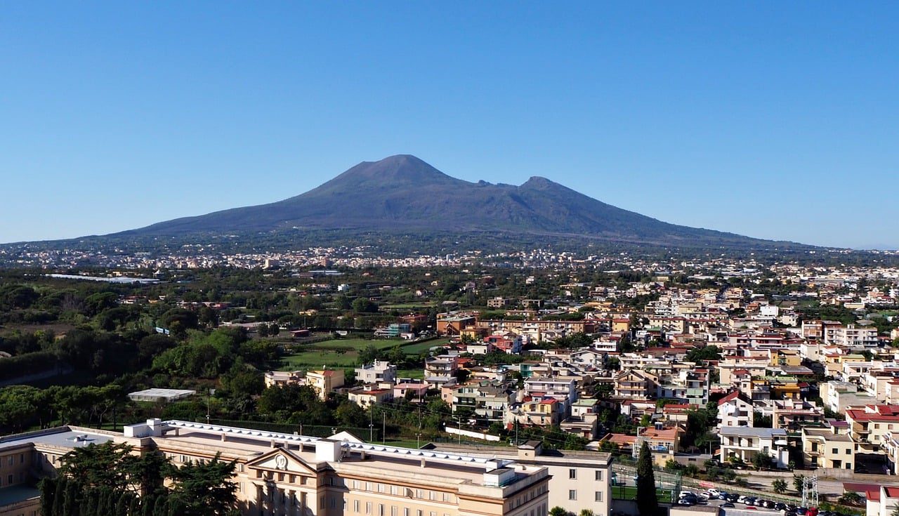 A imagem mostra uma vista panorâmica do Monte Vesúvio, com seu imponente cume vulcânico ao fundo, cercado por uma vasta área urbana. A cidade se estende pela planície, com casas, edifícios e vegetação espalhados, enquanto o céu está limpo, destacando o contraste entre a modernidade da cidade e a natureza poderosa do vulcão.