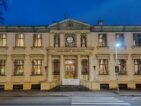 Fachada do Museu Laikrodziu em estilo neoclássico, um edifício histórico branco de dois andares iluminado à noite. A construção apresenta janelas ornamentadas com grades decorativas, estátuas na entrada principal, um vitral circular no centro e detalhes arquitetônicos elaborados. O nome do museu está inscrito na fachada em letras maiúsculas. Uma faixa de pedestres passa em frente ao edifício, que é iluminado por um poste de luz.