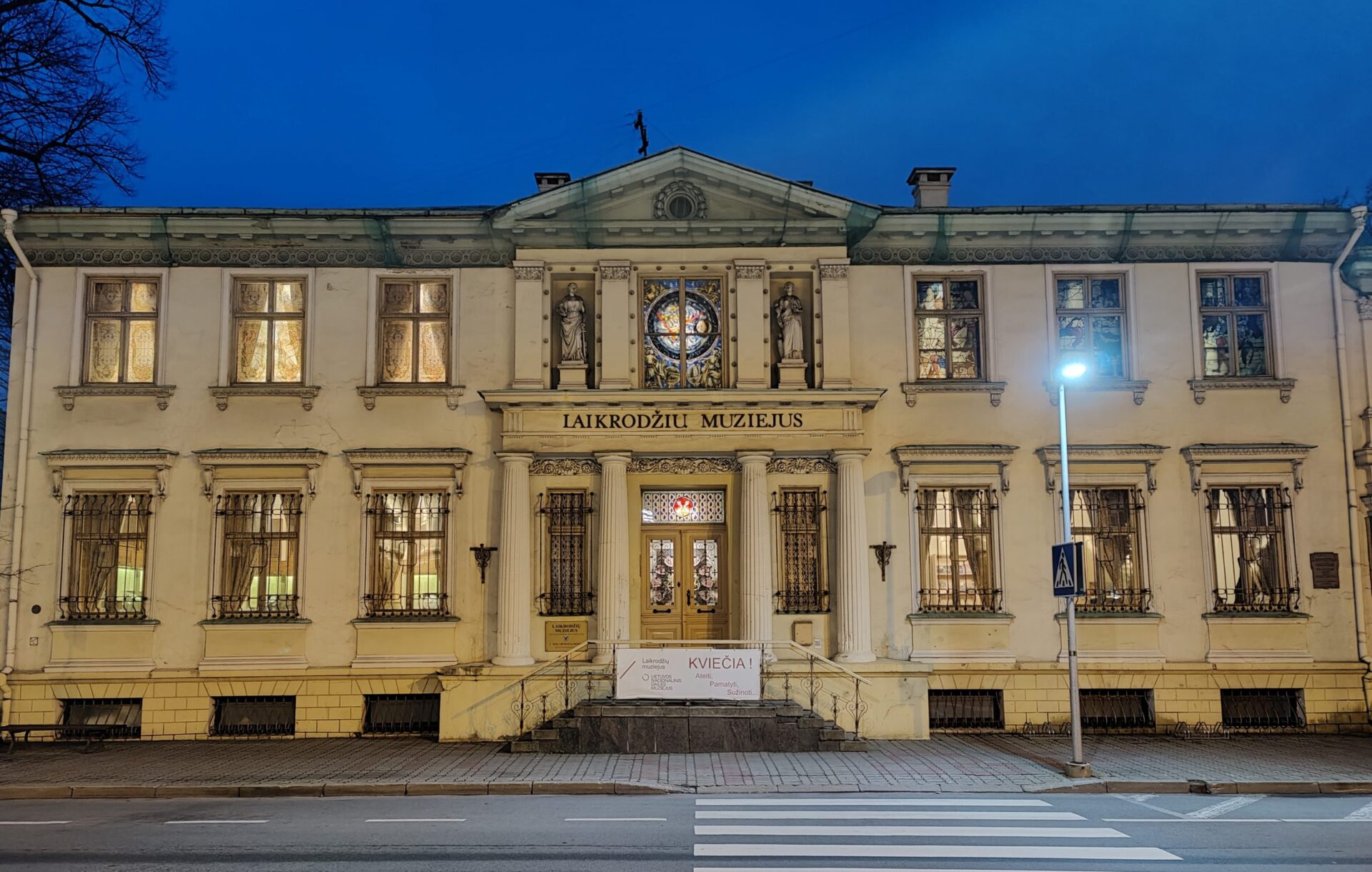 Fachada do Museu Laikrodziu em estilo neoclássico, um edifício histórico branco de dois andares iluminado à noite. A construção apresenta janelas ornamentadas com grades decorativas, estátuas na entrada principal, um vitral circular no centro e detalhes arquitetônicos elaborados. O nome do museu está inscrito na fachada em letras maiúsculas. Uma faixa de pedestres passa em frente ao edifício, que é iluminado por um poste de luz.