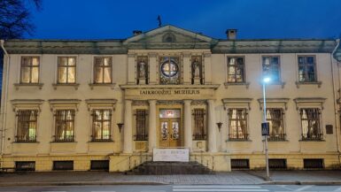 Fachada do Museu Laikrodziu em estilo neoclássico, um edifício histórico branco de dois andares iluminado à noite. A construção apresenta janelas ornamentadas com grades decorativas, estátuas na entrada principal, um vitral circular no centro e detalhes arquitetônicos elaborados. O nome do museu está inscrito na fachada em letras maiúsculas. Uma faixa de pedestres passa em frente ao edifício, que é iluminado por um poste de luz.