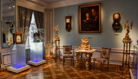 Sala elegante de museu exibindo relógios antigos e peças históricas em vitrines iluminadas, com móveis barrocos ornamentados, pintura clássica e relógios de parede decorativos contra paredes azul-claras. Piso em parquete e cortinas de época completam a atmosfera histórica.