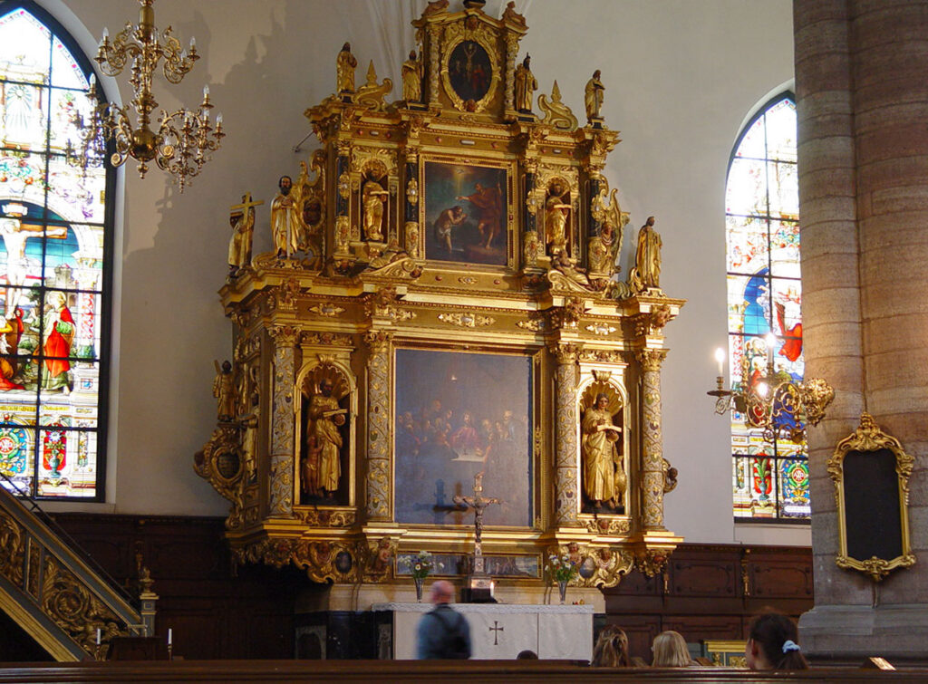 Interior da Igreja Alemã (Tyska Kyrkan) em Estocolmo, com um altar ricamente decorado em dourado, ladeado por estátuas e pinturas religiosas. Ao fundo, belos vitrais coloridos complementam a atmosfera reverente e artística do local.