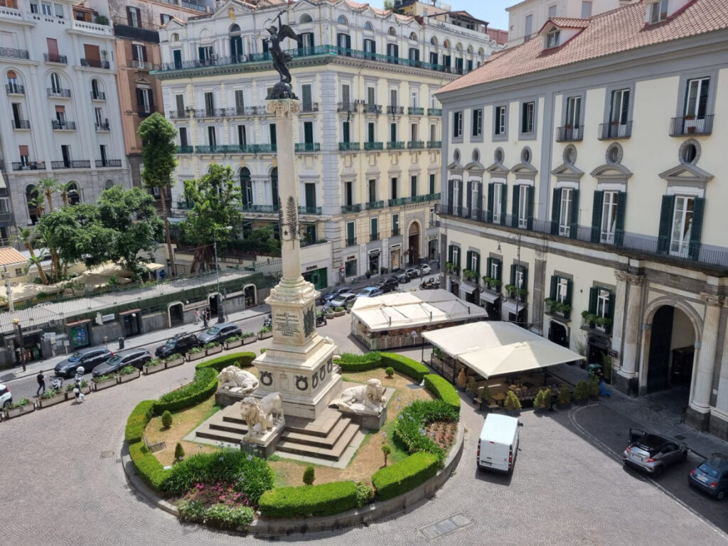 Vista aérea da Piazza dei Martiri, em Nápoles, destacando o monumento central com a coluna de mármore, esculturas de leões e áreas verdes ao redor. Cercada por edifícios históricos e cafés, a praça é um símbolo das lutas pela independência italiana.