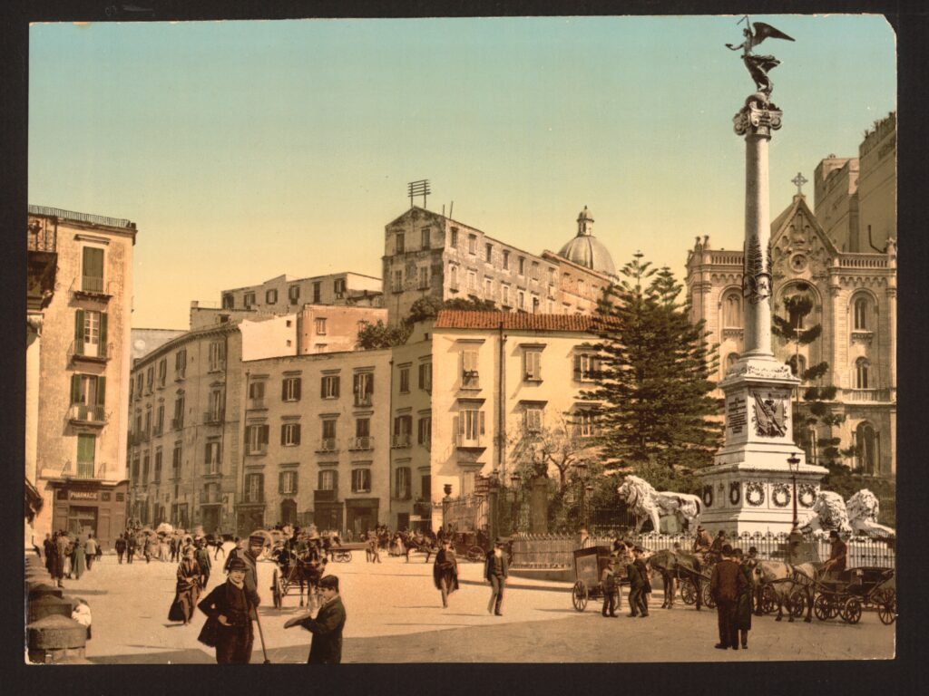 Imagem histórica da Piazza dei Martiri, em Nápoles, destacando o monumento central com a coluna e os leões esculpidos, cercado por edifícios clássicos e o movimento urbano do século XIX. Um símbolo da memória e elegância napolitana.