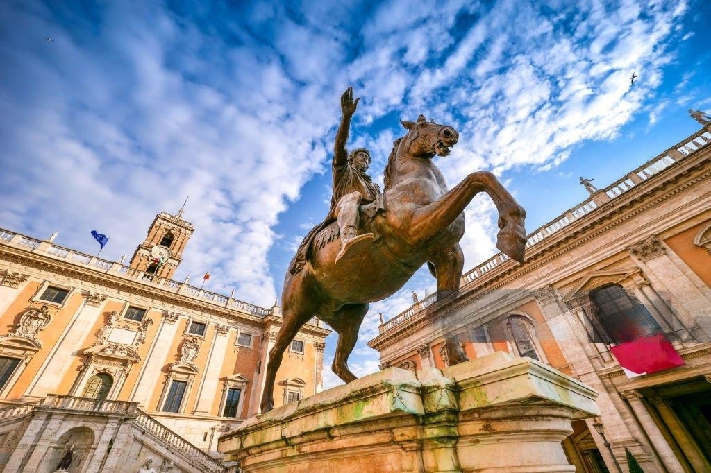 A icônica estátua equestre de Marco Aurélio na Piazza del Campidoglio, com o imponente Palazzo Senatorio ao fundo e o céu azul em contraste. A cena reflete a harmonia renascentista projetada por Michelangelo, combinando arquitetura, história e arte em um dos locais mais emblemáticos de Roma.