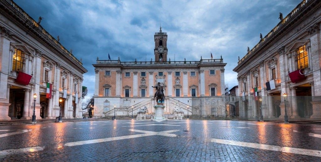 Vista ampla da Piazza del Campidoglio, com a elegante geometria renascentista de Michelangelo, destacando o Palazzo Senatorio ao centro, ladeado pelo Palazzo dei Conservatori e Palazzo Nuovo. O piso com padrões geométricos e o céu dramático completam a cena histórica.