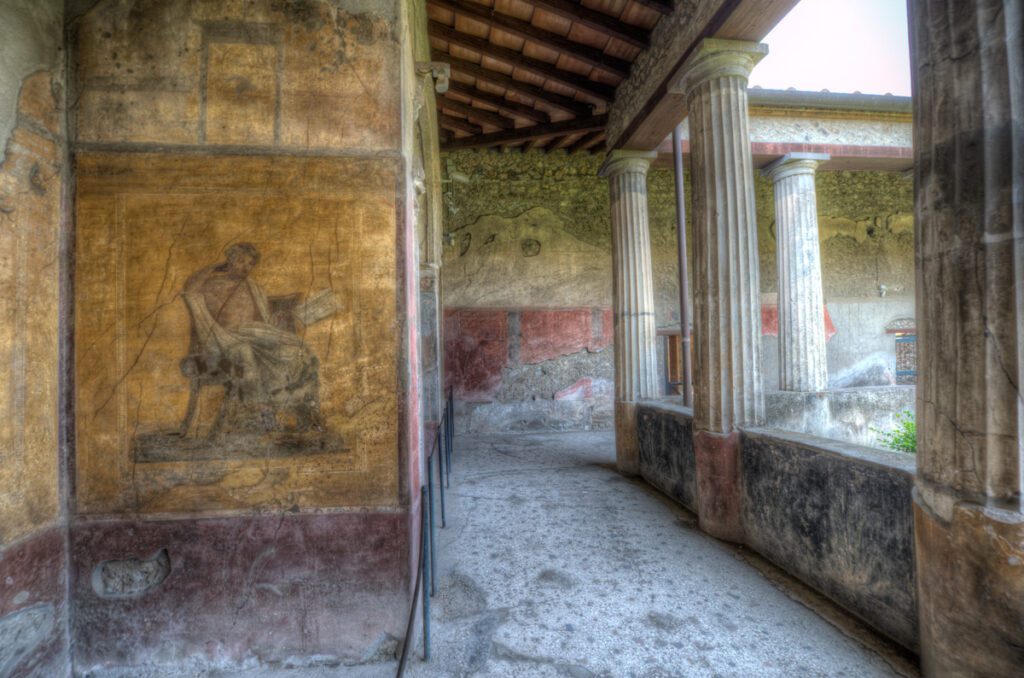Interior de uma casa romana em Pompeia, com colunas clássicas e um afresco retratando uma figura sentada, preservando detalhes arquitetônicos e artísticos da Antiguidade.
