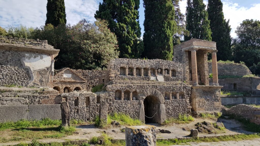 Ruínas romanas em Pompeia, com paredes de tijolos e detalhes arquitetônicos preservados, cercadas por vegetação verde e árvores altas.