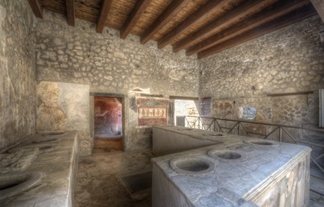 Interior de uma cozinha romana em Pompeia, com bancadas de pedra e afrescos nas paredes, preservando detalhes da arquitetura e da vida cotidiana da Antiguidade.