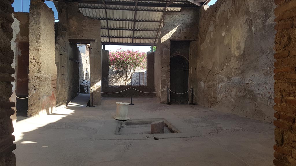 Interior de uma estrutura romana em Pompeia, com paredes de tijolos e um telhado metálico moderno, exibindo detalhes arquitetônicos preservados e vegetação ao fundo.
