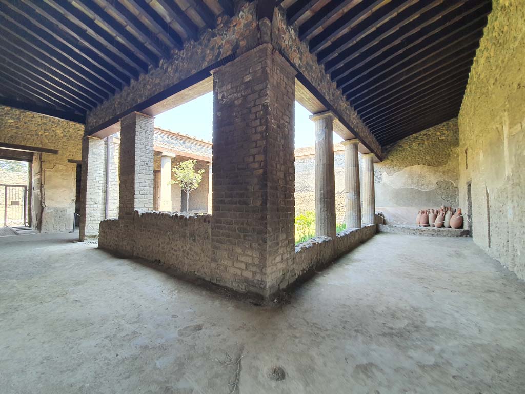 Interior de uma estrutura romana em Pompeia, com colunas e um teto de madeira, exibindo detalhes arquitetônicos preservados e vasos antigos no fundo.