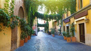 Uma rua tranquila em Roma, com paredes de tijolos e plantas pendentes formando um túnel verde sobre o pavimento de pedras. Um cenário charmoso que reflete a beleza tradicional da cidade.